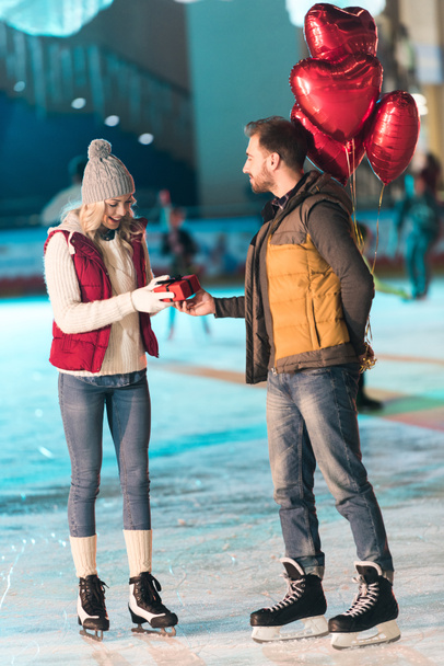 side view of young man with balloons presenting gift box to girlfriend at st valentines day on rink   - Foto, immagini