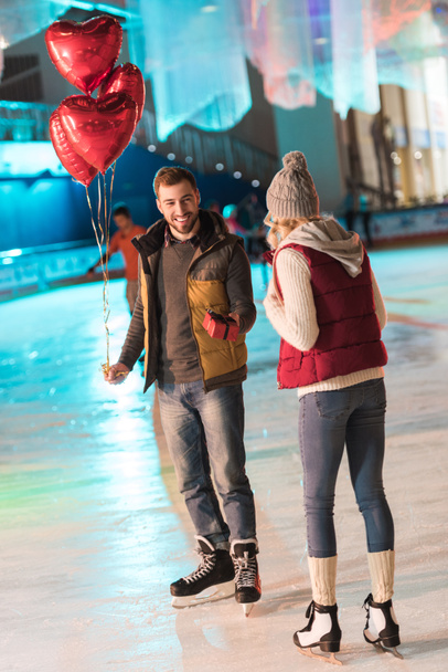 веселый молодой человек с воздушными шарами, подаривший подарочную коробку своей девушке на катке в День Святого Валентина
   - Фото, изображение