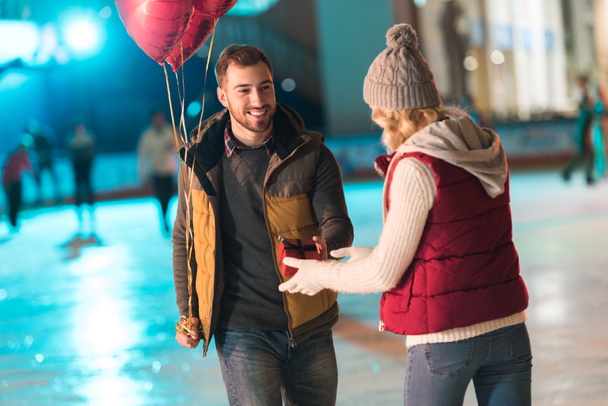 χαρούμενος νεαρός με μπαλόνια παρουσιάζοντας πλαίσιο δώρο στη φίλη στο st ημέρα του Αγίου Βαλεντίνου στο παγοδρόμιο   - Φωτογραφία, εικόνα
