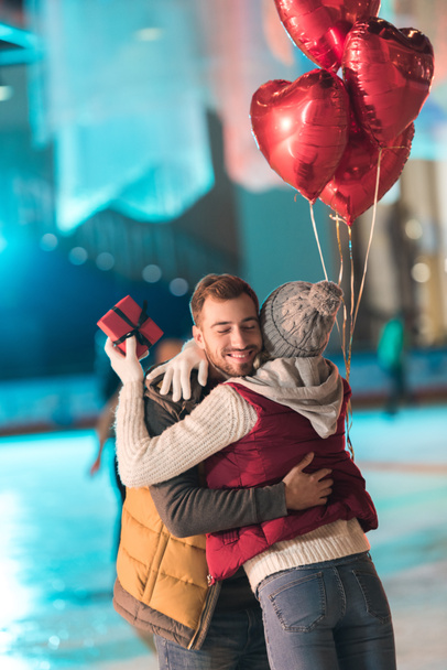 jovem mulher segurando caixa de presente e abraçando namorado feliz com balões em forma de coração na pista
  - Foto, Imagem
