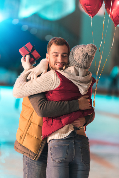 young woman holding gift box and hugging boyfriend with balloons on skating rink  - Photo, Image