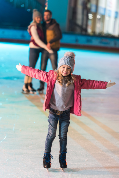 adorable petite fille debout avec les bras ouverts et souriant à la caméra tandis que les parents debout derrière sur la patinoire
 - Photo, image