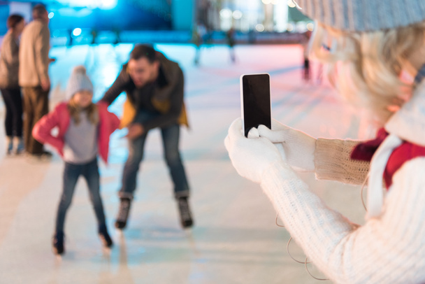 cropped shot of woman holding smartphone with blank screen and photographing family skating on rink  - Foto, imagen