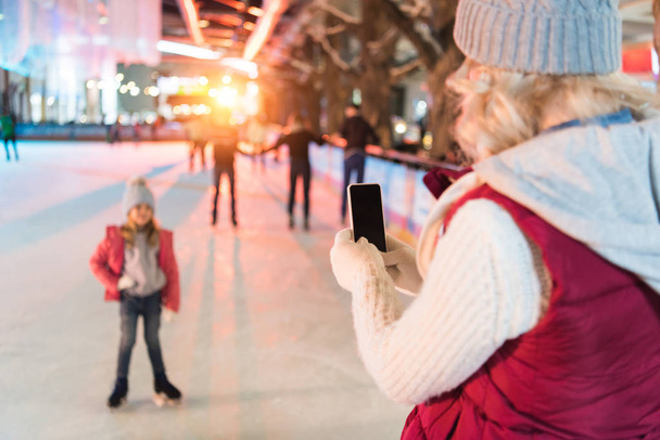 mujer joven con teléfono inteligente fotografiando linda hija pequeña en pista de patinaje
 - Foto, imagen