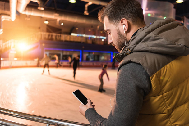 όμορφος νεαρός άνδρας στα ακουστικά χρησιμοποιώντας το smartphone με την κενή οθόνη για παγοδρόμιο - Φωτογραφία, εικόνα