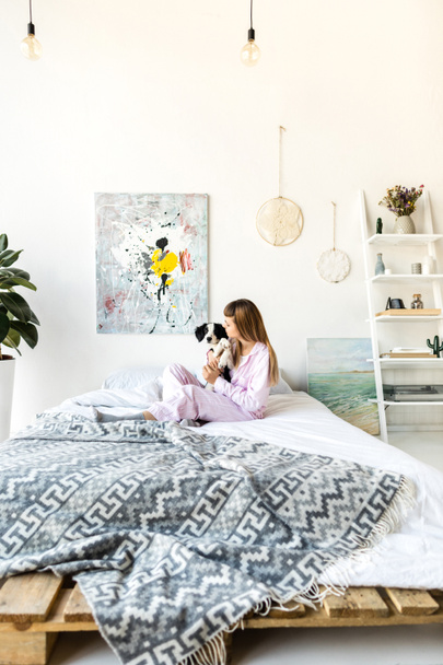 Młoda kobieta w piżamie przytulanie szczeniak siedząc na łóżku rano w domu - Zdjęcie, obraz