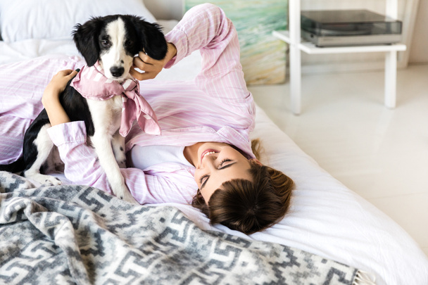 黒と白の子犬と一緒にベッドに横になっているパジャマの若い女性 - 写真・画像