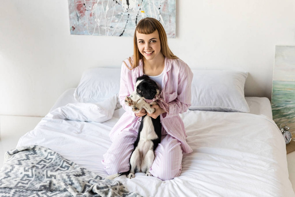 счастливая женщина в пижаме и милый маленький щенок отдыхает на кровати утром дома
 - Фото, изображение