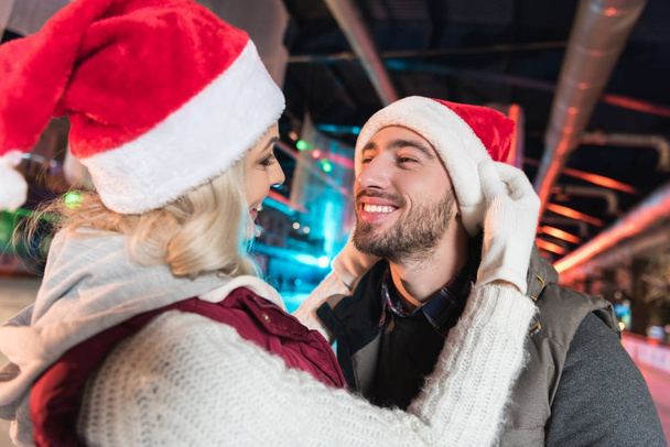 glückliches junges Paar in Weihnachtsmannhüten, die sich auf der Eisbahn gegenseitig anlächeln - Foto, Bild