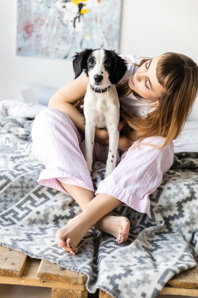 young woman hugging puppy while sitting on bed in morning at home - Foto, afbeelding