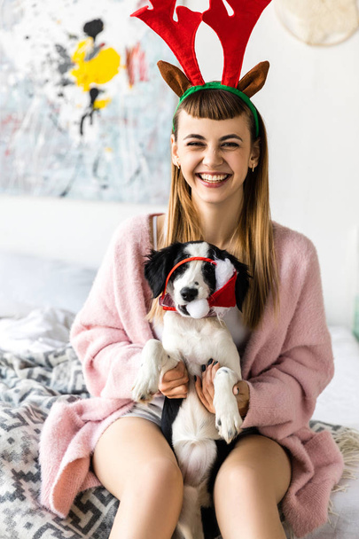 portrait of smiling woman with deer horns on head and puppy in santa hat  - Photo, Image