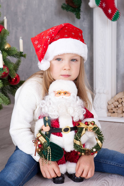Christmas or New year celebration. Little cute girl in santa hat on the head embraces a toy Santa Claus and sits near Christmas tree with xmas gifts. A fireplace with christmas stocking on background - Photo, Image