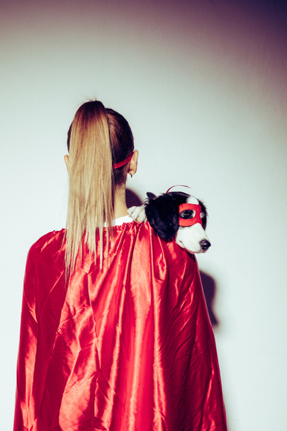 back view of woman in red cape holding little puppy in superhero mask - Foto, imagen