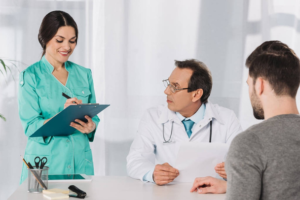 doctor looking at smiling nurse writing something to clipboard - Photo, Image