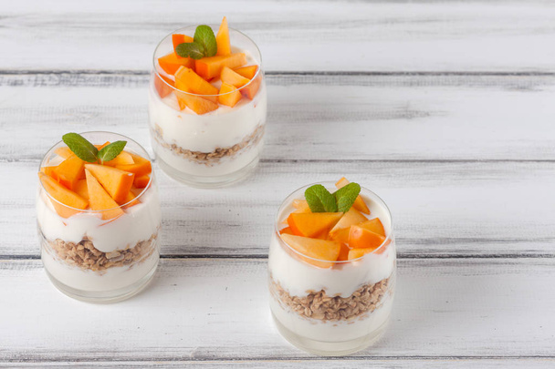 Persimmon creamy trifle in beautiful glasses, fresh ripe fruit slices on white wooden background. Healthy vegetarian food. Delicious dessert. Close up photography. Selective focus. - Photo, Image