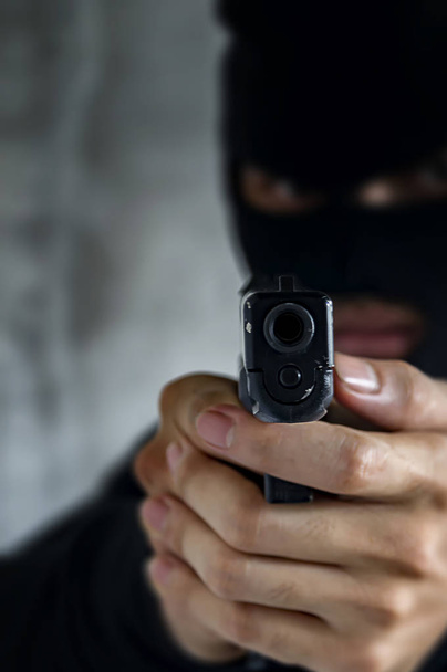 Ladrão mascarado com arma apontando para a câmera contra um ba preto
 - Foto, Imagem