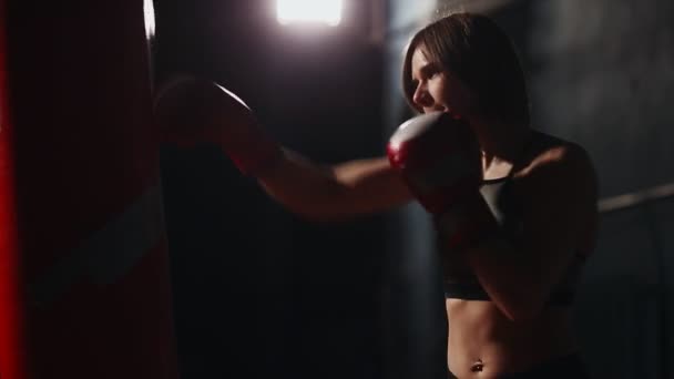 Sportieve brunette boksen sportschool biedt individuele training op stroke techniek. Voert strikes op een bokszak op een zwarte achtergrond. - Video
