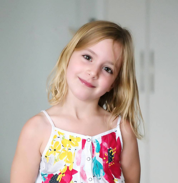 Симпатичная шестилетняя блондинка в белом платье с цветами, улыбающимися на абстрактном фоне
 - Фото, изображение