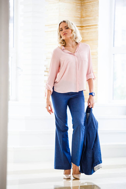 Portrait of beauty young blond woman with curly hair, wearing blue casual suit and pink shirt standing by window at home interior - Фото, изображение