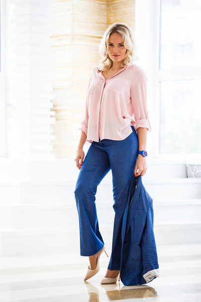 Portrait of beauty young blond woman with curly hair, wearing blue casual suit and pink shirt standing by window at home interior - Photo, Image