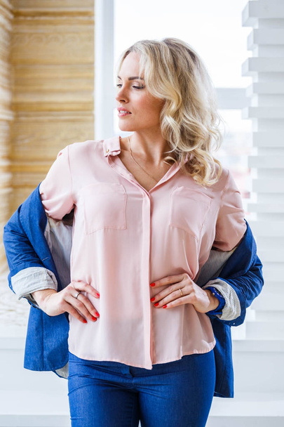 Portrait of beauty young blond woman with curly hair, wearing blue casual suit and pink shirt standing by window at home interior - Photo, Image