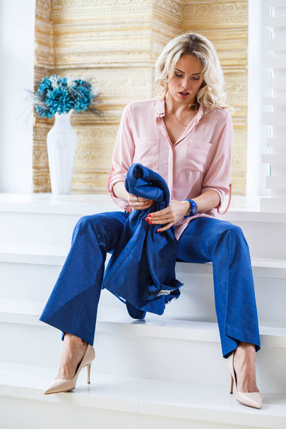 Portrait of beauty young blond woman with curly hair, wearing blue casual suit and pink shirt sitting on white stairs by column near window at home interior - Photo, Image