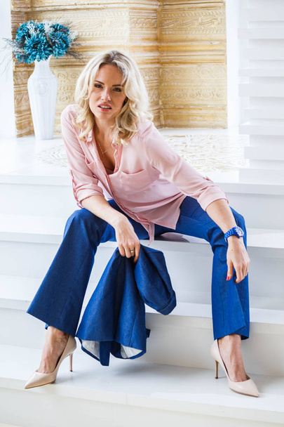 Portrait of beauty young blond woman with curly hair, wearing blue casual suit and pink shirt sitting on white stairs by column near window at home interior - Фото, зображення