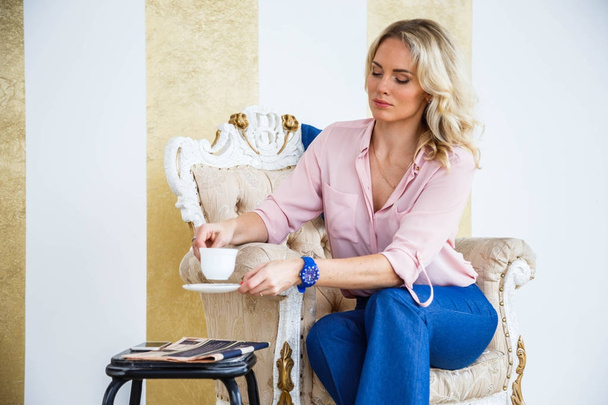 Portrait of beauty young blond woman with curly hair, wearing blue casual suit and pink shirt sitting in vintage armchair and drinking morning coffee. White and gold wall and floor at home interior background - Fotografie, Obrázek
