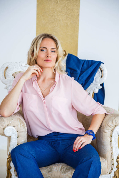 Portrait of beauty young blond woman with curly hair, wearing blue casual suit and pink shirt sitting in vintage armchair. White and gold wall and floor at home interior background - Photo, Image