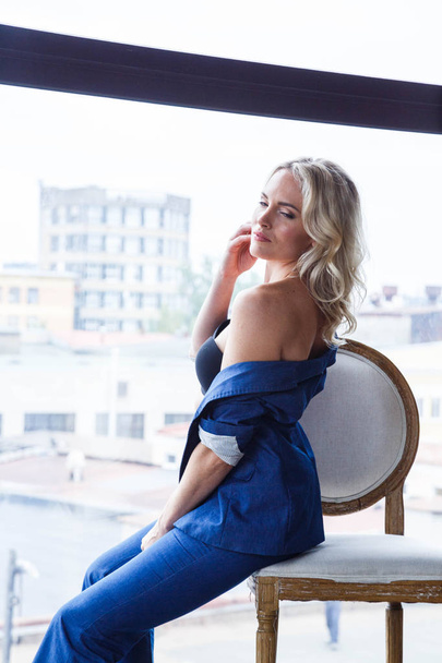 Studio portrait of beautiful blond woman with curly hair and stylish makeup, wearing blue casual suit and blue shoes on high hills, sitting on white vintage chair, blurred city view in window on background  - Photo, image