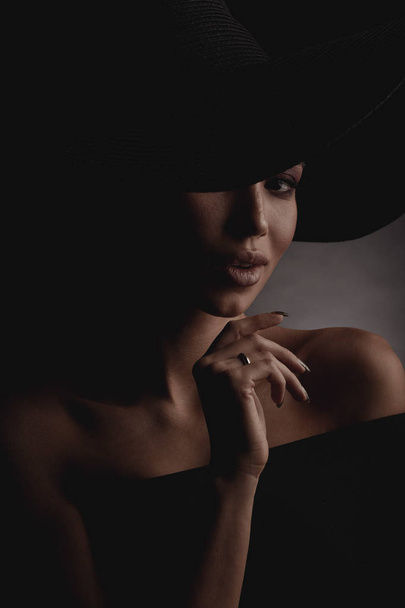 Dark studio portrait of elegant and sexy young woman in black wide hat and black dress.  - Photo, Image