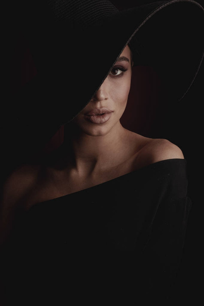 Dramatic dark studio portrait of elegant and sexy young woman in black wide hat and black dress.  - Φωτογραφία, εικόνα