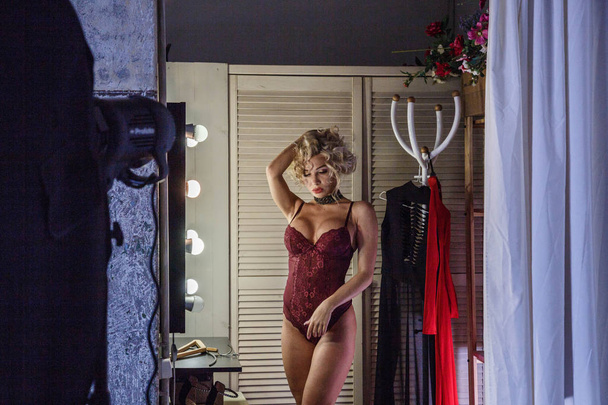 Beautiful young woman with curly blond hair, wearing beauty purple lingerie, standing near mirror illuminated by light-bulbs in wardrobe, wooden doors and red dress on background.  - Фото, изображение