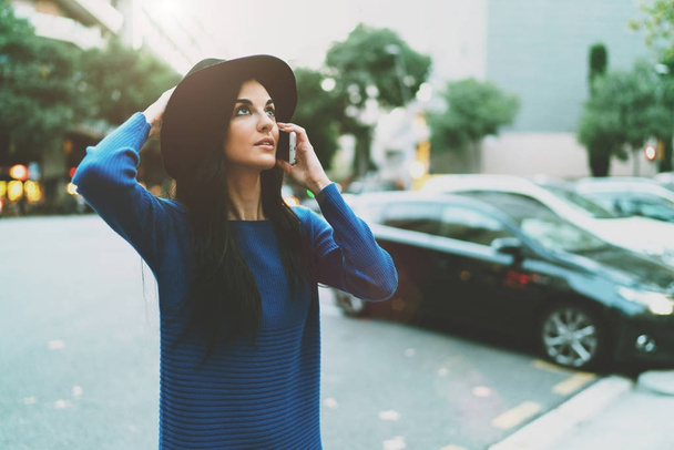 Charming student girl wearing stylish clothes is talking with a friend by a smartphone while standing on a blurred city background. Talented designer calling to the colleague while standing outdoors. - Foto, immagini