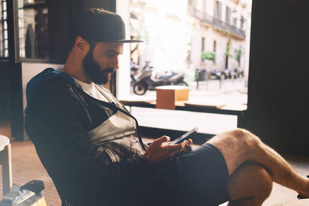 A bearded male is checking e-mail on a smartphone while sitting in a city coffee shop. Hipster guy wearing a snapback is surfing the web on a mobile phone. Handsome guy is texting by a cellular phone. - Foto, imagen