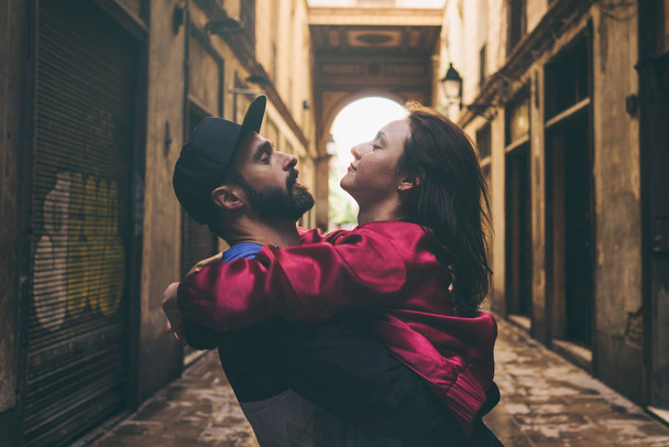A bearded man in a cap is hugging his beloved woman while standing on a street background. Attractive woman is looking into the eyes of her husband during spending a leisure time outdoors. - Photo, image