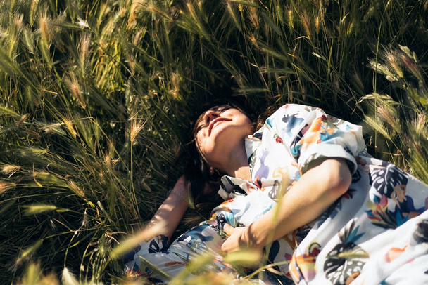 Portrait of an attractive caucasian female with dark hair looking at the skies while lying amidst the field. A model look woman in a colorful dress is relaxing outdoors on a sunny day. - Photo, image