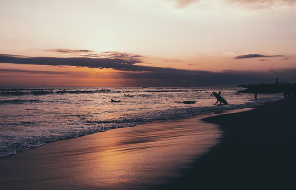 A silhouette of a surfer with a surfing board walking out the ocean on a dusk. Colorful sunset on a sandy beach of the tropical sea. Atmospheric photo of a nightfall on the ocean. - Foto, Imagem