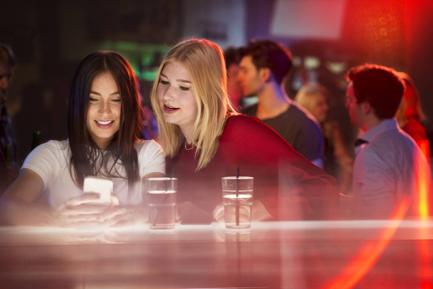 Two young attractive girls looking at smartphone at bar and laughin - Foto, Bild