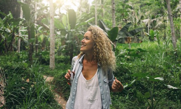 Happy tourist girl is exploring wild jungles in exotic country. Beautiful caucasian female with curly hair is impressed with exotic wild nature while visiting national park of tropical plants. - Фото, зображення