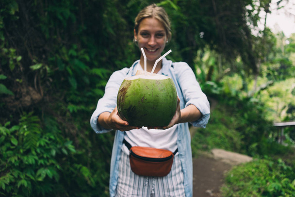 Young woman with blonde hair is offering a coconut cocktail on a green jungle background. Green coconut with straws in hands of a cheerful girl walking the tropical forest during summer vacation. - Foto, Imagen