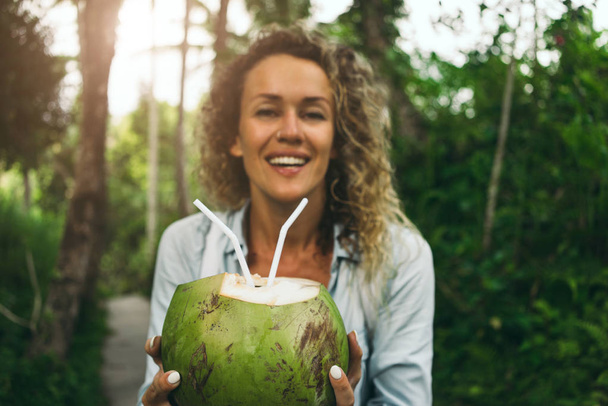 Young smiley woman with curly hair is offering a coconut cocktail on a green jungle background. Green coconut with straws in hands of a happy girl walking the tropical forest during summer vacation. - Фото, изображение