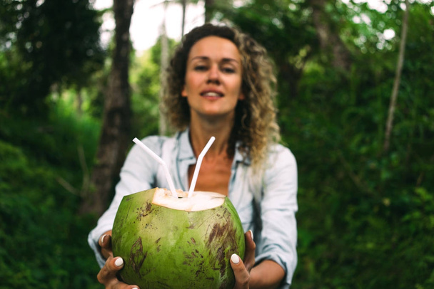 Young woman with curly hair is offering a coconut cocktail on a green jungle background. Green coconut with straws in hands of a tourist female walking the tropical forest during summer vacation. - 写真・画像