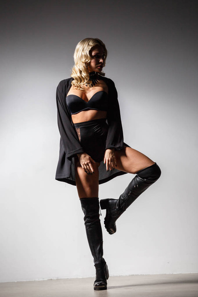 Full-length studio portrait of beautiful blond woman wearing black clothes and black boots standing on grey wall background  - Photo, image
