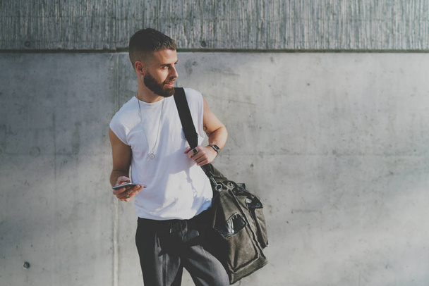 Smiling bearded model look guy in casual clothes with empty area for logo or design  chatting with friend via social network on a mobile phone. Handsome caucasian male with a smartphone in his hand - Zdjęcie, obraz