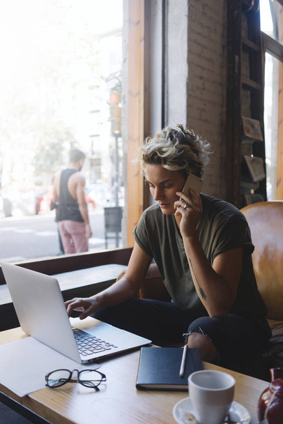 Caucasian female is talking by a mobile phone while having rest in a coffee shop. Beautiful blonde woman is surfing the web on a portable computer while relaxing with a cup of coffee. - 写真・画像