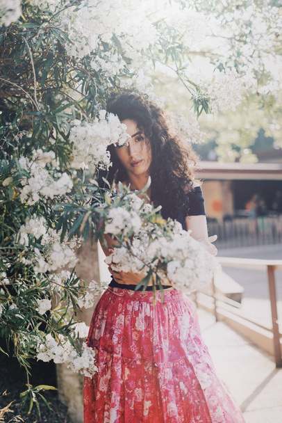 Young caucasian female wearing colorful summer skirt and black top is looking at the camera while standing beside the flowers. A brunette woman with long curly hair is having rest outdoors. - Fotó, kép