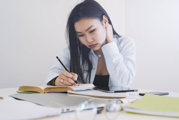 International student girl is working on a diploma project while sitting at the table with a laptop, digital tablet, smartphone and books on it. Young asian freelancer is working at home. - 写真・画像