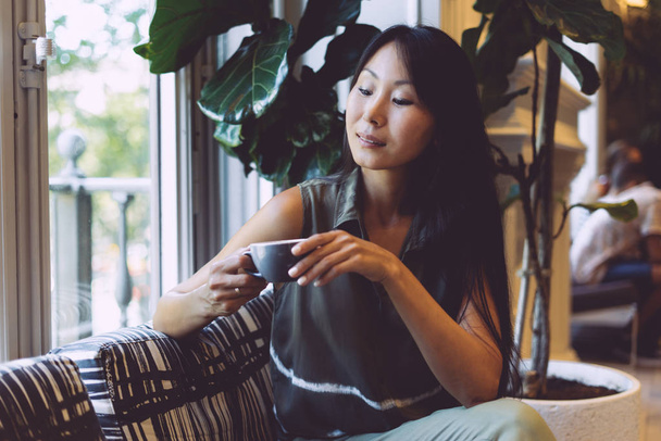 Portrait of a beautiful asian woman with long dark hair holding a cup of coffee in a modern coffee shop. Trendy fashion designer is drinking tea on a blurred background of a city cafe. - Foto, imagen