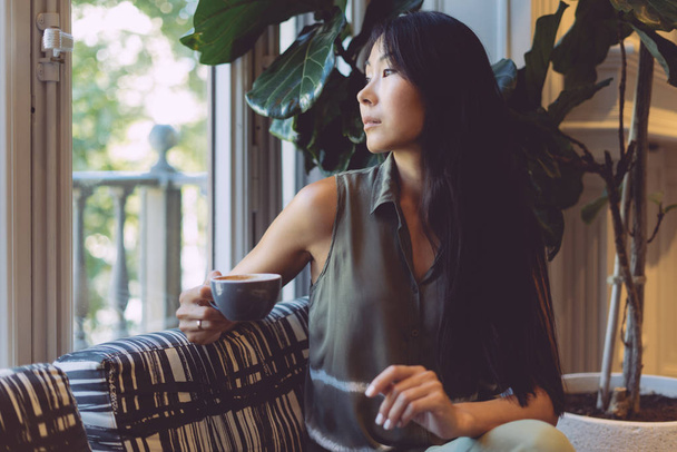 Uroczy dojrzale kobiet o długich ciemnych włosach jest korzystających herbaty rano siedząc w przytulnej kawiarni. Portret uroda blogger ubranie, siedząc z filiżanką kawy w café noszenia. - Zdjęcie, obraz